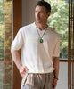 Man wearing a white polo shirt and beige pants with a green jade peace buckle pendant necklace, standing indoors.