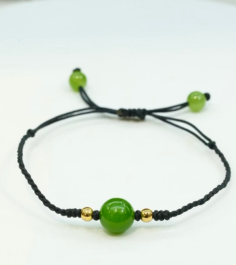 Close-up product view of an apple green, AAA grade natural Siberian Hetian jade bead bracelet featuring a vibrant nephrite jade centerpiece, small gold-filled accent beads, and an adjustable black cord band.
