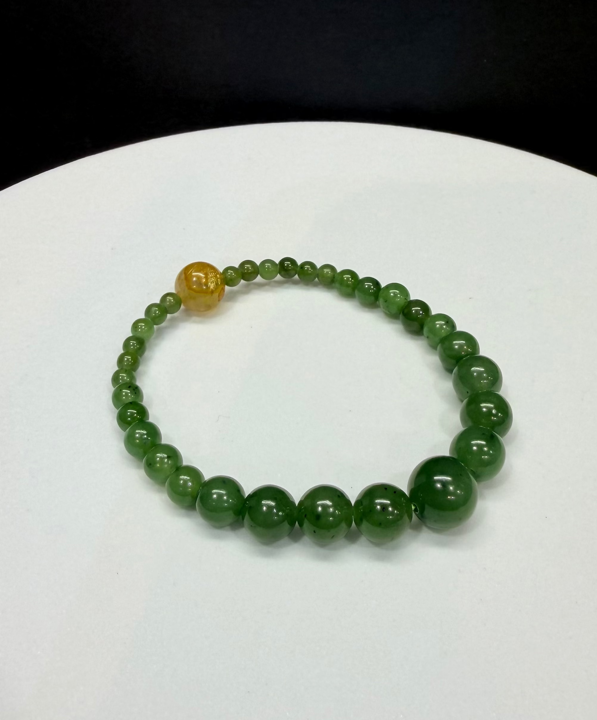 Green arctic nephrite jade beaded bracelet with descending bead sizes and a single gold rutilated quartz crystal bead on a white background.
