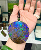 A person holding a large, round Canadian Ammolite pendant featuring vibrant, iridescent shades of electric blue, green, and purple with a distinct natural crackle pattern.