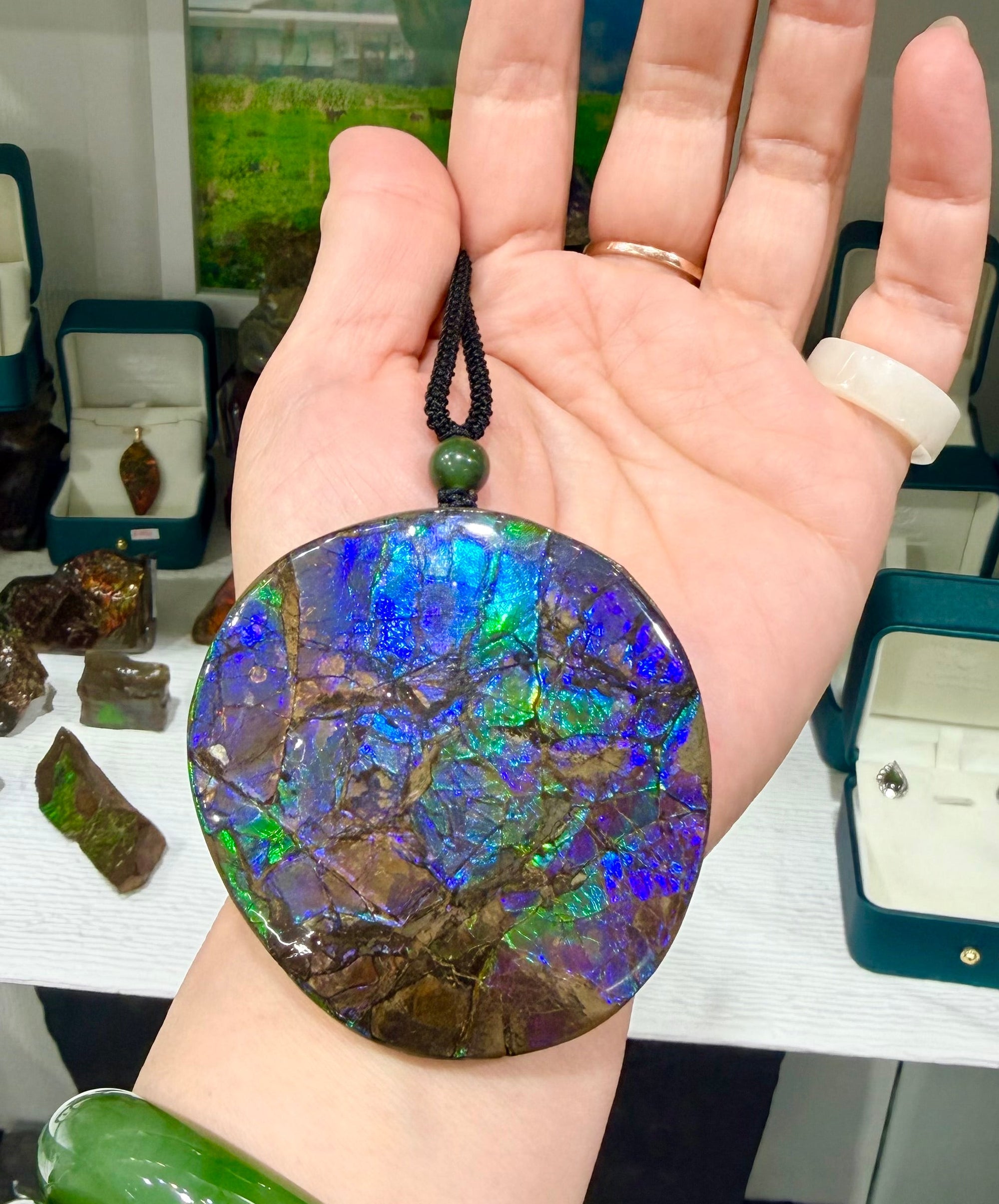 A person holding a large, round Canadian Ammolite pendant featuring vibrant, iridescent shades of electric blue, green, and purple with a distinct natural crackle pattern.