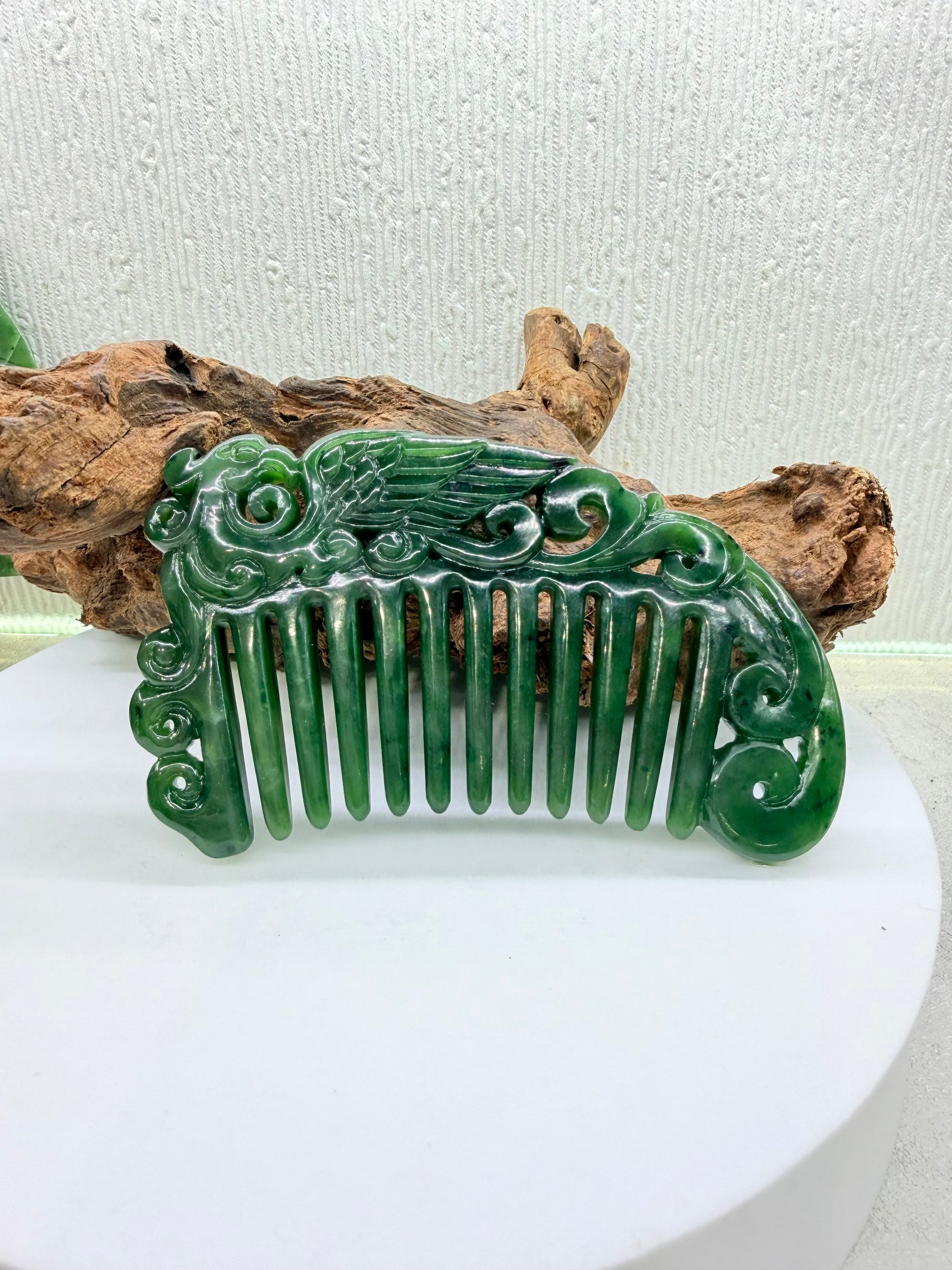 Green nephrite polar phoenix wing jade comb on a white pedestal with wooden logs in the background