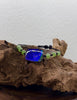 A beaded ammolite jade bracelet with a central displayed on a piece of driftwood against a textured white wall.