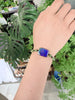 A person's wrist wearing a beaded bracelet with a central rectangular purple ammolite stone, surrounded by green Jade beads, set against a background of green houseplants.