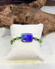 A beaded bracelet with a central iridescent blue-purple rectangular stone ammolite and green round Jade beads, displayed on a white cushion in front of a piece of driftwood.